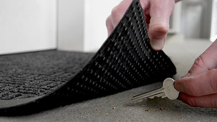 Zwei Hände heben eine Fußmatte an, darunter liegt ein Schlüssel