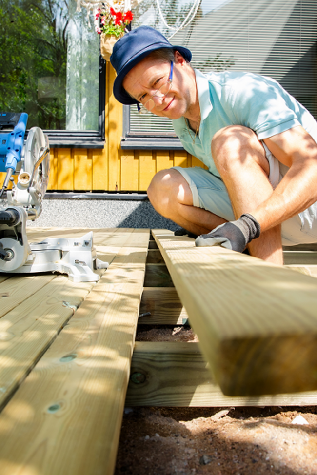 Mann baut Holzterrasse vor seinem Tiny House und schaut dabei in die Kamerla